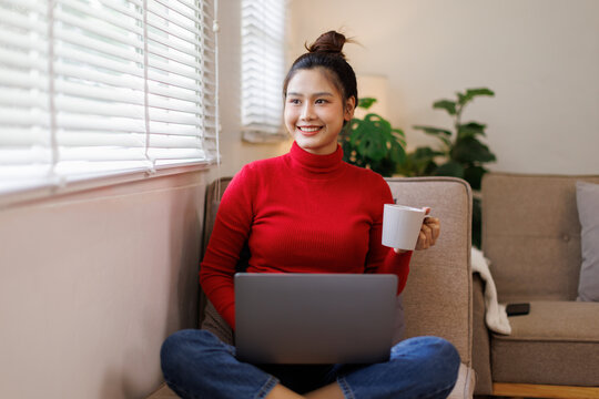 Relax Concept. Happy woman drinking coffee using pc sitting on comfortable couch at home in living room and dreaming. Cheerful casual famale resting on sofa, enjoying weekend free time.
