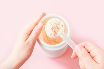 Female hand holding measuring spoon with dry vanilla protein powder on pink background.