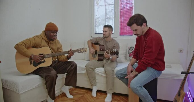 Three friends enjoy making music together at home. Two young men play guitars while their friend plays the cajon and sings.