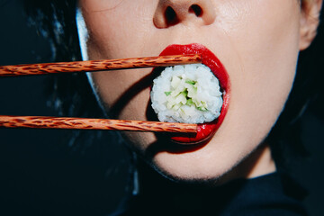 Close-up of a woman with bold red lips holding sushi in her mouth with chopsticks, highlighting the...
