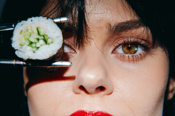 Close-up portrait of a woman holding sushi with chopsticks, showcasing vibrant eyes and bold red...