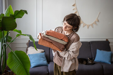 Serene woman bringing clean fresh laundry in living room. Bedding refresh, clothing hygiene,...