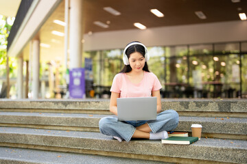 Young asian student girl smiling happy wearing headphone using laptop  while sitting on stairs...