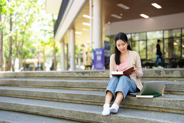 Asian college student sitting on the stairs of a university campus, reading a book and studying....