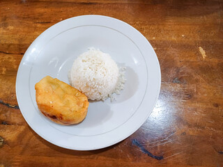 White plate holding a scoop of white rice and a piece of fried bakwan, presented on a wooden table....