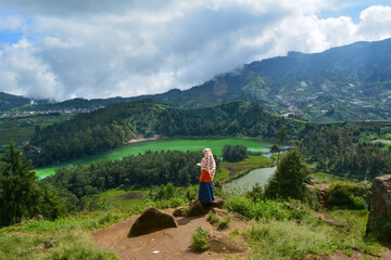 A girl in a hijab standing on a hilltop, overlooking the scenic Dieng Plateau with its lush green...
