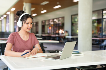 Smiling young asian woman in wireless headphones sitting at table in cafeteria working on laptop...
