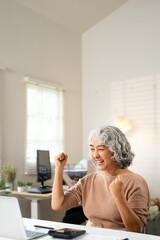 Happy senior woman raising arms in excitement while using laptop at home, celebrating online win,...