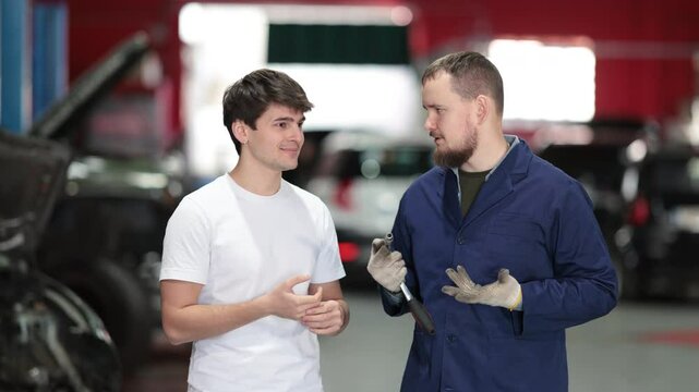 Young customer of a car service communicates about the process of repairing a car with a qualified mechanic. Specialist gives recommendations on oil changes, engine checks and tire changes