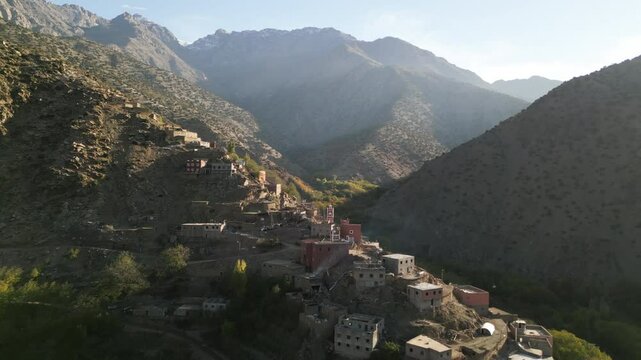 Drone shot of a sunlit Berber village on a hillside in the High Atlas mountains in Morocco, surrounded by autumn trees and dramatic peaks on a clear morning
