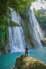 Nature traveler faces tall waterfalls cascading into calm blue waters, framed by rocks and forest vegetation