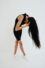 slender woman with long black hair in black sleeveless outfit bending forward against plain white...