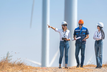 Three people are standing on a dirt road, looking at a large wind turbine
