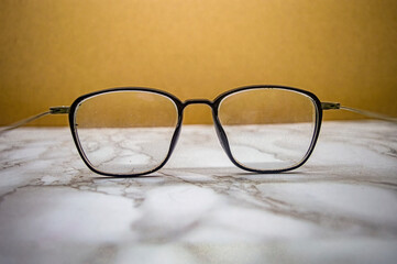 Black square eyeglasses with subtle dust on clear lenses placed over marble surface and beige backdrop, combining everyday realism with minimalist fashion and vision clarity