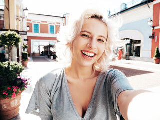 Young beautiful smiling hipster woman in trendy summer clothes. Sexy carefree model posing on the street background at sunset. Positive blond female outdoors. Cheerful and happy. Taking Pov selfie