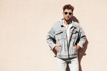 Portrait of handsome confident stylish hipster model. Sexy man dressed in jeans jacket. Fashion male posing in the street. In sunglasses in Europe city. Outdoors at sunset.  Near wall