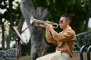 Musician practices trumpet while reading sheet music on a bench under the trees