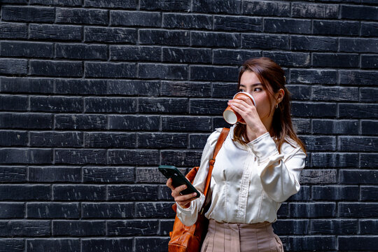 Businesswoman drinking takeaway coffee and using her smartphone in front of a black brick wall