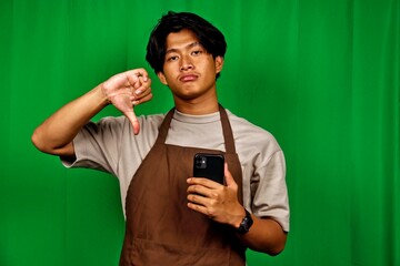 Young man wearing a beige shirt and brown apron holding a smartphone with a thumbs down gesture...