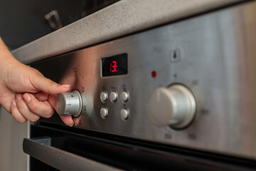 Close-up of a woman's hand adjusting a silver control knob on a modern built-in kitchen oven with a...