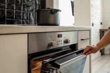 Close-up of a hand opening a hot oven in a modern kitchen. The oven is glowing inside, ready for...