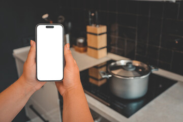 Two hands holding smartphone with blank screen in modern kitchen. Cooking pot on induction stove in...