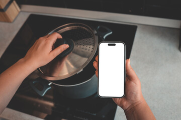 Person holding smartphone with blank screen in one hand while lifting pot lid with the other over...
