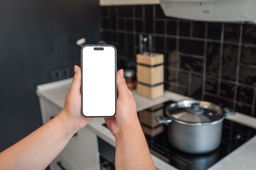 Two hands holding smartphone with blank screen in modern kitchen. Cooking pot on induction stove in...