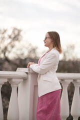Balcony Woman Pink Outfit: Woman in white blazer looks pensively from balcony on a cloudy day...
