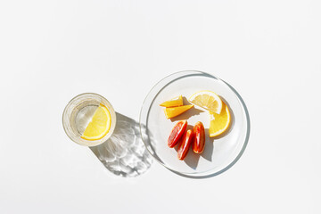 Summer still life scene with clear glass cups and plate. Glittering glasses of water with slices of fruit on white table at sunlight. Long harsh shadows and caustics. Summer vacation concept, above