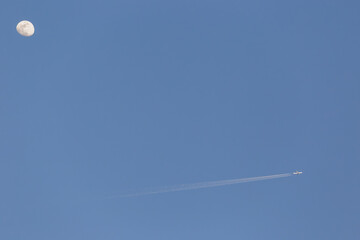 Commercial airplane flying across a clear blue sky, leaving a contrail, with the moon visible in...