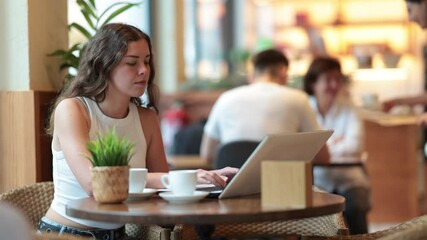 Young woman is sitting in a coffee shop with a laptop and is carefully typing. European woman works remotely from a cafe. Woman communicates on the Internet in social networks - Powered by Adobe
