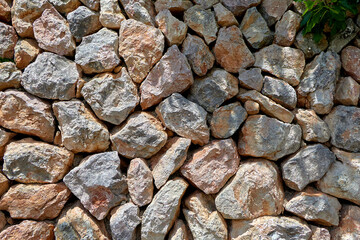 Rustic dry stone wall made of rough, irregular rocks in natural shades of gray, brown, and orange, tightly packed and sunlit. stone wall;