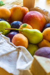 A beautifully arranged basket filled with a colorful mix of fresh fruits including red apples, green pears, dark plums, orange apricots, and nectarines. The basket is lined with a textured natural