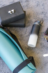 Flat lay composition of modern yoga and fitness essentials on a soft grey carpet. The image includes a rolled-up turquoise yoga mat with a black strap, a reusable water bottle with a gradient black