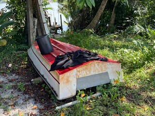 A weathered red boat lies overturned by the riverside, showing signs of rust and decay. Scattered items rest atop its corroded surface, hinting at abandonment and the passage of time