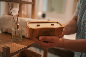Woman holding a handmade wooden candle with two wicks in a cozy bathroom. The scene includes a bamboo shelf, aroma diffuser, and soft towel, creating a relaxing, spa-like atmosphere