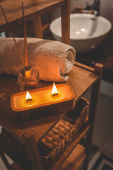 A handmade wooden candle with twin flames burning brightly on a bamboo shelf in a spa-style bathroom, accompanied by a rolled towel and glass diffuser, creating a serene and cozy atmosphere