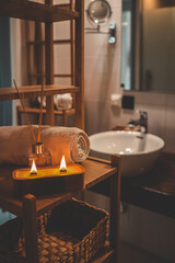 A handmade wooden candle with twin flames burning brightly on a bamboo shelf in a spa-style bathroom, accompanied by a rolled towel and glass diffuser, creating a serene and cozy atmosphere