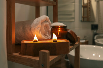 Burning wooden wick candle with amber jar and rolled towel on bamboo shelf in cozy bathroom. Warm...