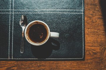 Overhead view of a white cup filled with dark coffee and a spoon on a black textured placemat over a wooden table. Morning light casts shadows, creating a warm and cozy coffee moment