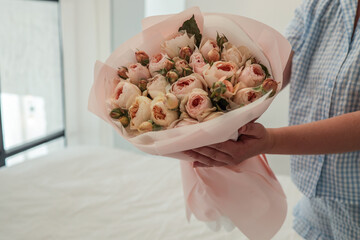 Close-up of a woman in a blue gingham outfit holding a bouquet of pastel roses wrapped in pink paper, set against a soft, minimal bedroom background with natural light