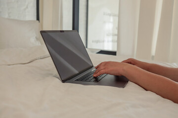 Woman working on a laptop while lying on a white bed in a bright, minimalist bedroom with sheer...