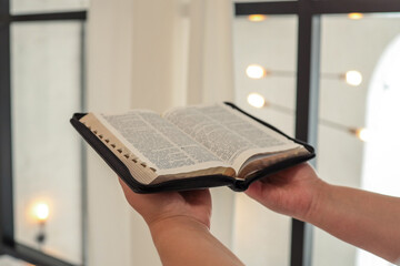 Hands holding an open Bible in a bright, modern interior with large windows and warm lighting,...