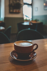 Warm and cozy coffee shop interior with a ceramic cup of cappuccino on a wooden table, soft lighting, blurred background with plants, and a relaxed, inviting atmosphere