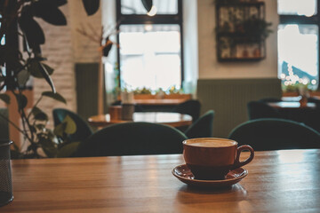Warm and cozy coffee shop interior with a ceramic cup of cappuccino on a wooden table, soft lighting, blurred background with plants, and a relaxed, inviting atmosphere