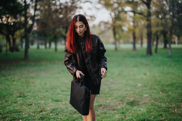 Young woman with red hair wearing a leather jacket and dress, standing outdoors in a park holding a...