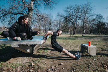 A man performing tricep dips with the help of a woman offering guidance in an outdoor park setting...