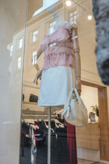 Mannequin in a pink ruffled blouse and white skirt holding a cream handbag, displayed in a boutique window with soft lighting and reflections of classic architecture outside