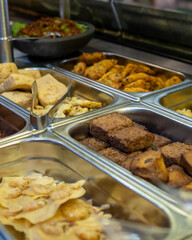 Assorted traditional Indonesian fried snacks such as tempeh, tofu, and fritters displayed in stainless steel food trays at a food stall or restaurant.